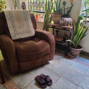 Image shows a vacant recliner and a pair of slippers on the floor in front of the recliner. The background features a lot of plants.