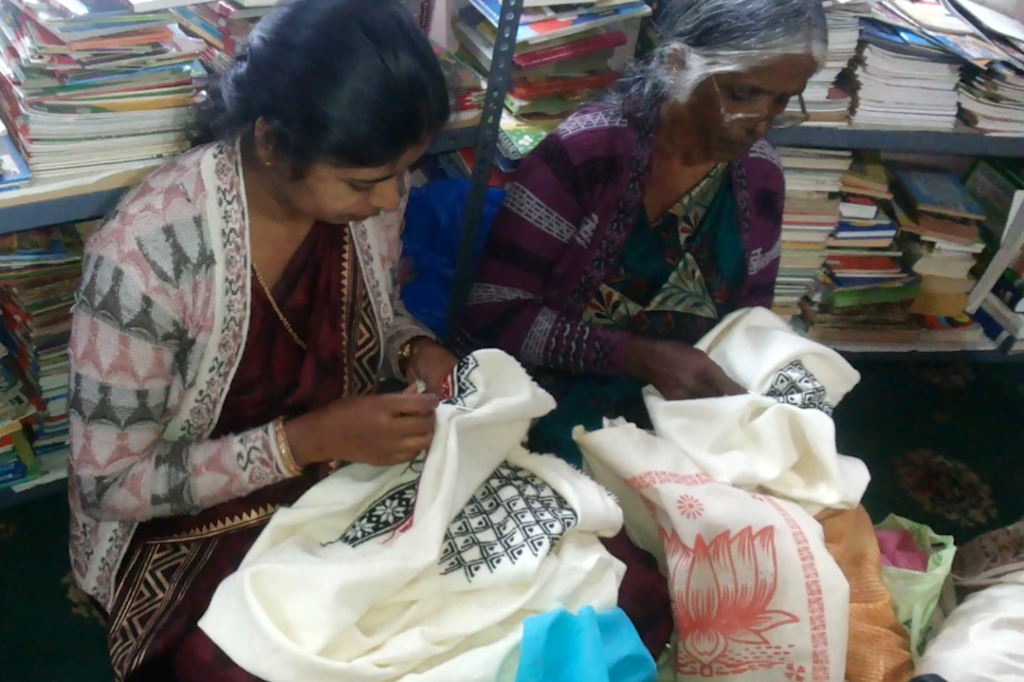 Two women sitting on the floor and creating Toda embroidery on white cloth