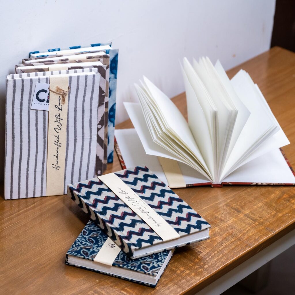 A set of diaries with blockprinted designs as the cover, with handmade paper, on a brown table top. One of the diaries is open. 