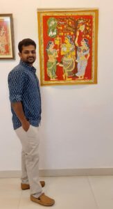 Artist Saikiran standing on the left and a cherial painting on the wall to the right of him. The painting depicts three women under a tree, engaged in beauty rituals.